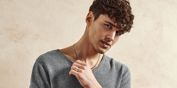 Junger Mann mit lockigen Haaren in grauem Strickpullover trägt silberne Panzerkette und silbernen Ring 